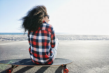 Hispanische Frau sitzt auf einem Skateboard am Strand - BLEF03299