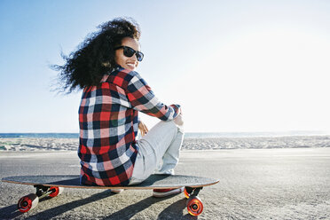 Hispanische Frau sitzt auf einem Skateboard am Strand - BLEF03300