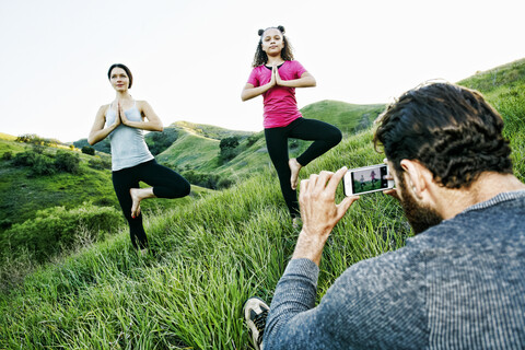 Man photographing wife and daughter practicing yoga on hill stock photo