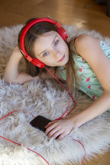 Mädchen liegt zu Hause auf dem Teppich und hört mit Kopfhörern und Smartphone Musik - SARF04281