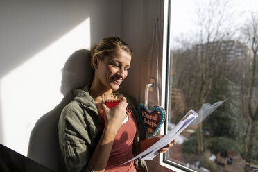 Lächelnde junge Frau, die am Fenster Zeitung liest - GUSF01960