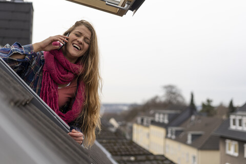 Glückliche junge Frau mit Handy, die sich aus dem Dachfenster lehnt, lizenzfreies Stockfoto