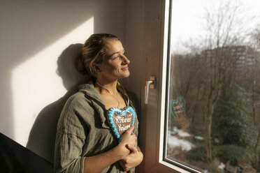 Lächelnde junge Frau am Fenster mit Lebkuchenherz - GUSF01967