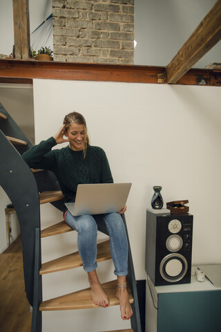 Lächelnde junge Frau sitzt auf einer Treppe zu Hause und benutzt einen Laptop, lizenzfreies Stockfoto