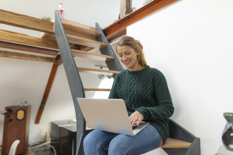 Lächelnde junge Frau sitzt auf einer Treppe zu Hause und benutzt einen Laptop, lizenzfreies Stockfoto