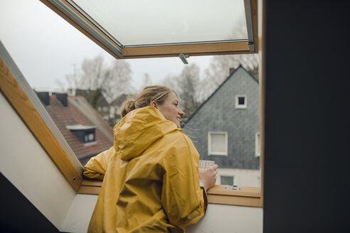 Lächelnde junge Frau im Regenmantel, die aus dem Dachbodenfenster schaut - GUSF01989