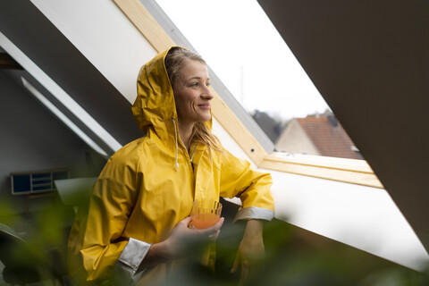 Lächelnde junge Frau im Regenmantel, die aus dem Dachbodenfenster schaut, lizenzfreies Stockfoto