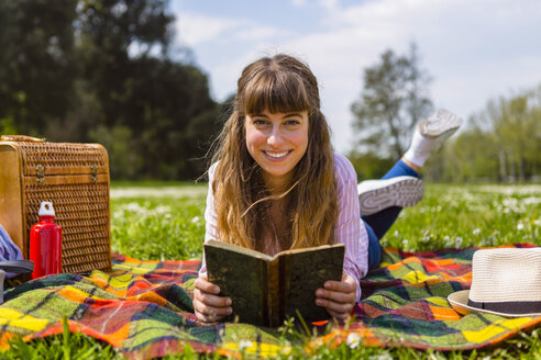 Junge Frau liegt auf einer Picknickdecke und liest ein Buch in einem Park - MGIF00471