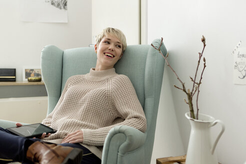 Happy woman sitting in armchair at home with tablet - FLLF00155