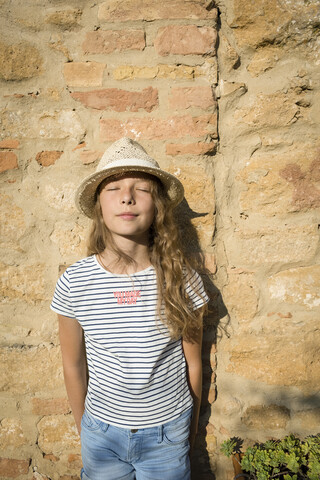 Mädchen mit Strohhut, das mit geschlossenen Augen an der Wand lehnt, Toskana, Italien, lizenzfreies Stockfoto