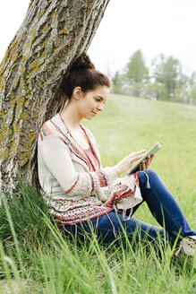 Junge Frau sitzt an einem Baum auf dem Land und benutzt ein Tablet - HMEF00368