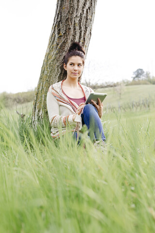 Junge Frau sitzt an einem Baum auf dem Land und benutzt ein Tablet, lizenzfreies Stockfoto