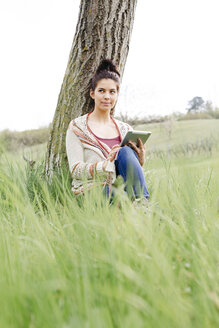 Junge Frau sitzt an einem Baum auf dem Land und benutzt ein Tablet - HMEF00369