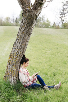 Junge Frau sitzt an einem Baum auf dem Land und benutzt ein Tablet - HMEF00370