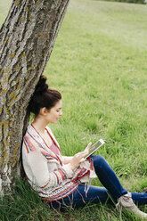Young woman sitting at a tree in the countryside using tablet - HMEF00371