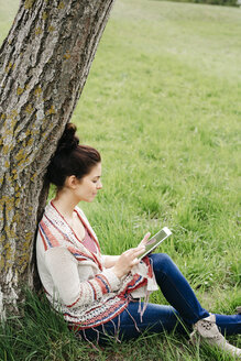 Junge Frau sitzt an einem Baum auf dem Land und benutzt ein Tablet - HMEF00371