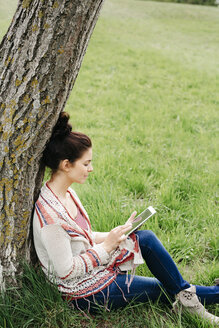 Young woman sitting at a tree in the countryside using tablet - HMEF00371