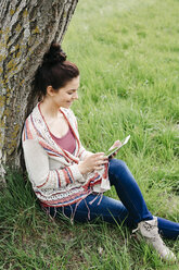 Lächelnde junge Frau, die an einem Baum auf dem Lande sitzt und ein Tablet benutzt - HMEF00372