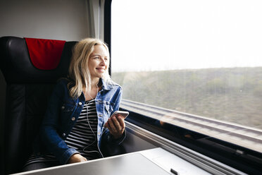 Glückliche blonde Frau, die mit Smartphone und Kopfhörern Musik hört und mit dem Zug reist - HMEF00376