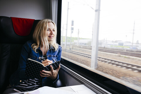 Porträt einer blonden Frau mit Notizbuch, die mit dem Zug reist und aus dem Fenster schaut, lizenzfreies Stockfoto