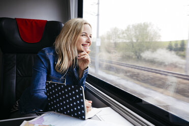 Lachende blonde Frau mit Notizbuch, die mit dem Zug reist und aus dem Fenster schaut - HMEF00380