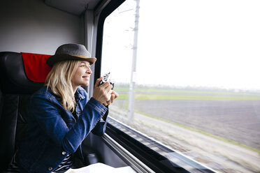 Glückliche blonde Frau mit Kamera, die im Zug reist und aus dem Fenster schaut - HMEF00382