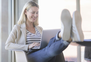 Lächelnde, entspannte Geschäftsfrau, die im Büro einen Laptop benutzt und die Füße hochlegt - UUF17480