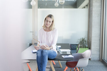Businesswoman sitting on desk in office using cell phone - UUF17486