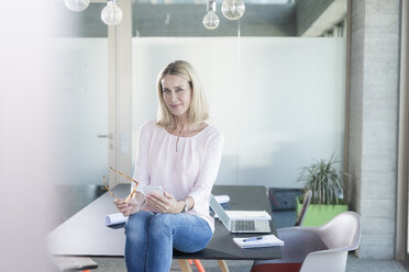 Portrait of confident businesswoman sitting on desk in office - UUF17487