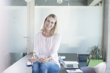 Portrait of smiling businesswoman sitting on desk in office - UUF17488
