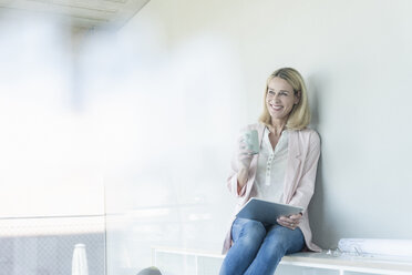 Happy businesswoman sitting on a shelf in office using tablet - UUF17496