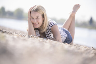 Lächelnde reife Frau auf Kieselsteinen am Flussufer liegend - UUF17598