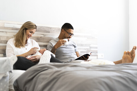 Mother bottle-feeding her newborn baby in bed with father reading book stock photo