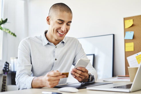 Junger Mann sitzt im Home Office und bezahlt online mit Kreditkarte und Smartphone - BSZF01075