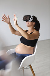 Junge schwangere Frau, die in einem Sessel sitzt und ihr Baby mit Hilfe einer Virtual-Reality-Brille beobachtet und fühlt - HMEF00385