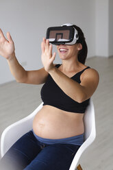 Junge schwangere Frau, die in einem Sessel sitzt und ihr Baby mit Hilfe einer Virtual-Reality-Brille beobachtet und fühlt - HMEF00386