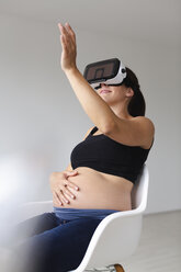 Junge schwangere Frau, die in einem Sessel sitzt und ihr Baby mit Hilfe einer Virtual-Reality-Brille beobachtet und fühlt - HMEF00387