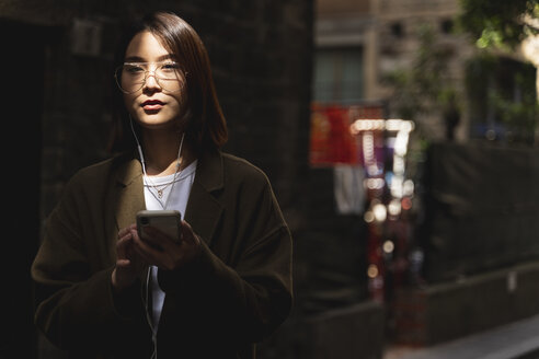 Italy, Florence, young woman with cell phone and earphones in sunlight in the city - FMOF00625