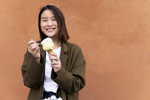Glückliche junge Frau isst eine Eistüte an einer orangefarbenen Wand - FMOF00632