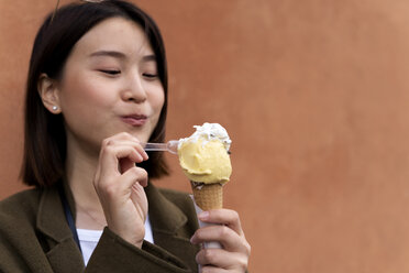 Porträt einer jungen Frau, die an einer orangefarbenen Wand eine Eistüte isst - FMOF00635