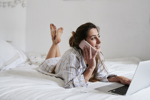 Junge Frau liegt im Bett, benutzt einen Laptop und telefoniert, lizenzfreies Stockfoto