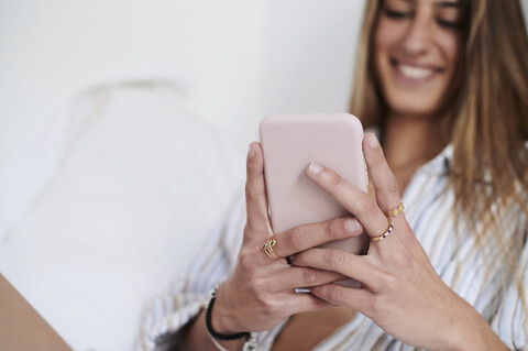 Junge Frau sitzt auf dem Bett und benutzt ihr Smartphone, lizenzfreies Stockfoto