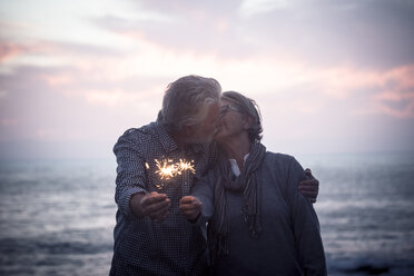 Kissing senior couple standing in front of the sea by sunset holding sparklers - SIPF01982