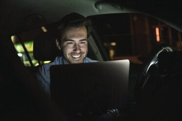 Lächelnder junger Mann mit Laptop im Auto bei Nacht - UUF17610