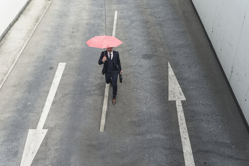 Geschäftsmann mit Regenschirm geht auf Straße mit Pfeil Zeichen - UUF17653