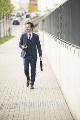 Businessman walking in the city with cell phone and earphones - UUF17672