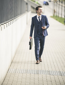 Happy businessman walking in the city with cell phone and earphones - UUF17673