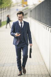 Businessman walking in the city with cell phone and earphones - UUF17675