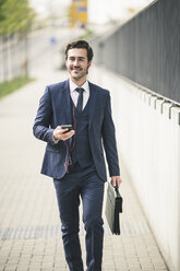 Businessman walking in the city with cell phone and earphones - UUF17676