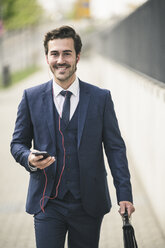 Businessman walking in the city with cell phone and earphones - UUF17677